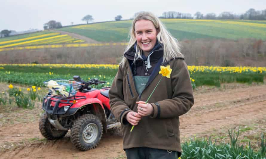 Frances Hosking standing next to a quad bike, smiling, holding a single daffodil, with a backdrop of yellow-tinged fields behind her