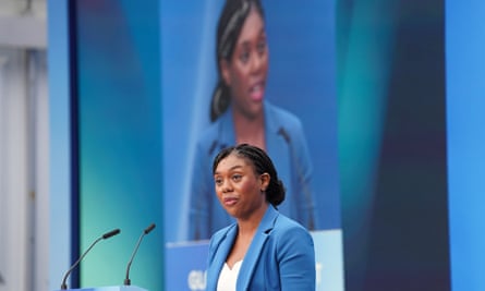 Kemi Badenocjh speaking at a podium at an event, with her image visible on a large screen behind her