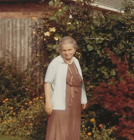 An elderly woman wearing a brown dress and a pale, short-sleeved cardigan, pearls and her glasses on a chain round her neck, standing in a garden with roses, other plants and a wooden fence behind her 