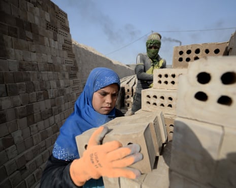 A girl wearing a blue headscarf and tradesperson's gloves that are too big for her adds a pile of bricks to a stack while a man watches her