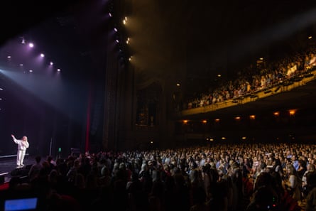 Robbins standing on stage at one of her sold-out events dressed in a white tracksuit, with a view of massive audience