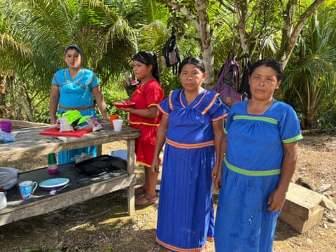 Four women in brightly coloured embroidered dresses stand at a wooden table with cups and plates on it, next to palm trees