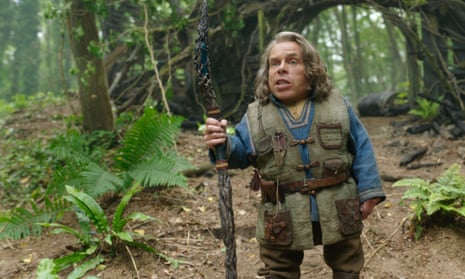 Warwick Davis, in a camouflage vest, holding a carved stick next to a fern in the forest