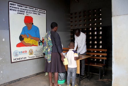 A mother and child stand in front of a medic’s desk near a mural bearing a USAID logo