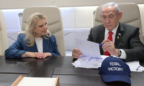 Benjamin Netanyahu sits at a table onboard a jet  looking at some official papers as his wife looks on. In front of Netanyahu is a blue cap with the words 'total victory' on it