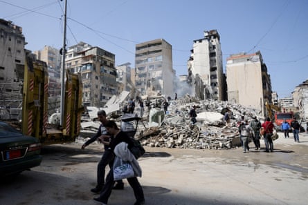 People carrying baggage walk past piles of debris in front of a destroyed apartment block.