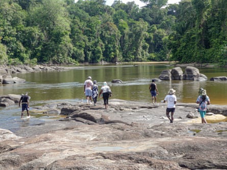 Seven people walk on an expanse of rock exposed by low water in a wide river bordered by jungle