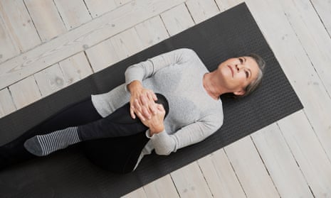 A woman with short hair wearing a grey long-sleeved  top and black leggings is lying on her back and stretching on a black yoga mat