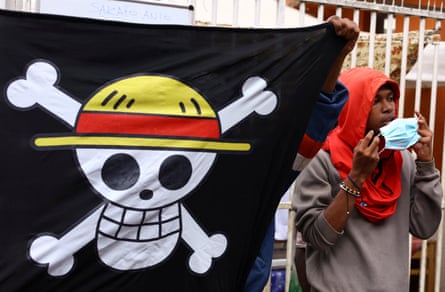 Protester puts on mask while another holds up a flag of a skull (wearing a red and yellow hat) and cross bones.