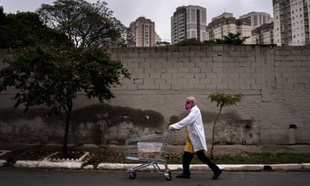 An elderly man in a facemask, white lab coat and yellow apron pushes a shopping trolley of food