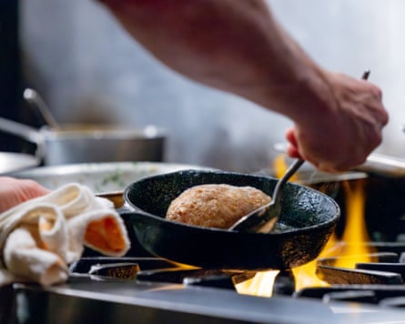 In the middle of the frame a cast iron skillet sizzles with a duck breast. A chef’s arm and hand reach into frame and prod the food with a spatula. Fire comes up from the cooktop below.