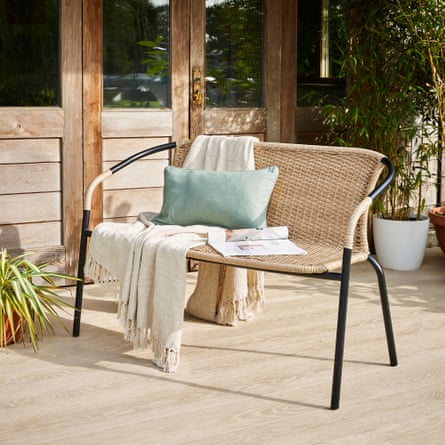 A rattan bench with metal frame sits on an outside decking with a cushion, throw and magazine laid out on the bench.