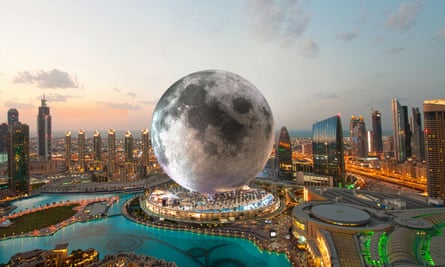 A giant model of the moon sits on a pedestal surrounded by palm trees and a lagoon, with Dubai skyscrapers arrayed behind it