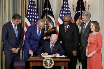 a man in a suit signs a document while seated at a table surrounded by people standing