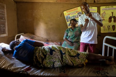 A man stands over a woman lying on a bed. Another woman sits on another bed behind him