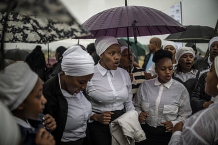 Group of mainly women, some with umbrellas, sing