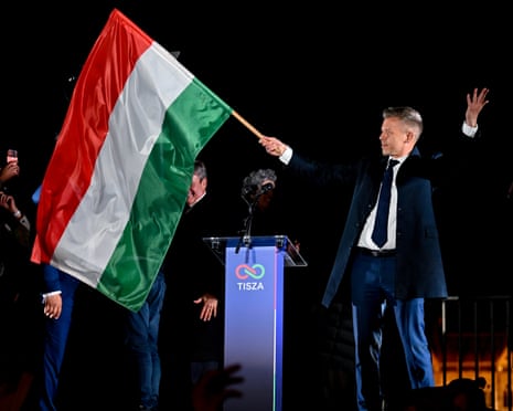 Magyar waves flag with one hand, his other hand in the air, beside podium, with illuminated building in the background
