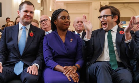 Kemi Badenoch, sitting next to Robert Jenrick on one side and her husband, Hamish, on the other, who is jubilantly clapping and wildly grinning.