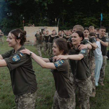 they are in a line with their hands on one another’s shoulders as if in a conga. They are casually dressed in short-sleeved khaki shirts with badges on the sleeves and camouflage combat trousers. The girls have their hair tied back.