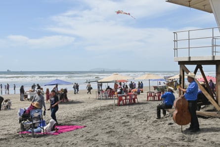 A sandy beach with dozens of people sitting or walking along it, a pink towel, and someone playing a standup bass.