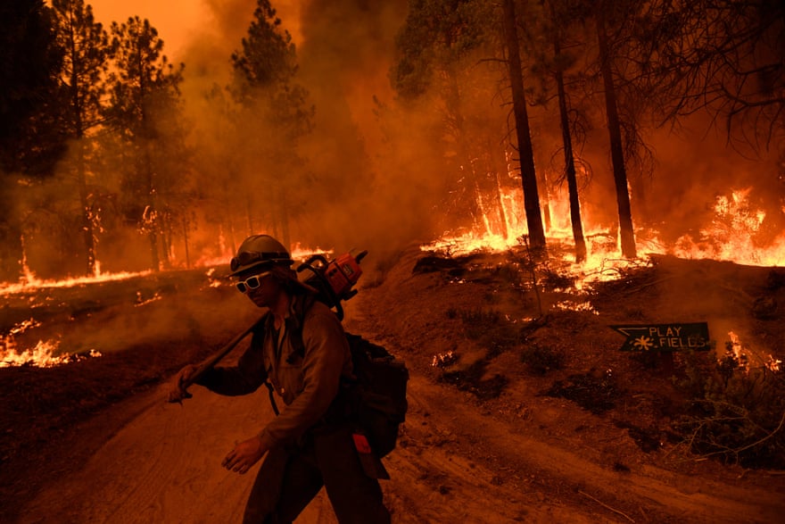 A firefighter wielding a chainsaw is surrounded by flames as a forest fire turns the forest orange and fills it with smoke.