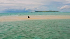 Photo of man sitting on sandbar in water with expanse of water in foreground and background. An island of land can be seen on the horizon