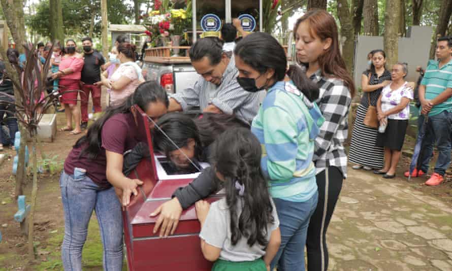 Young women and a man lean over a red coffin crying, while other people stand around solemnly