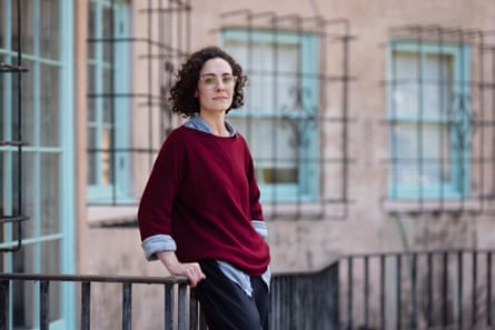 A woman in a red sweater leans against a black fence against the backdrop of a building
