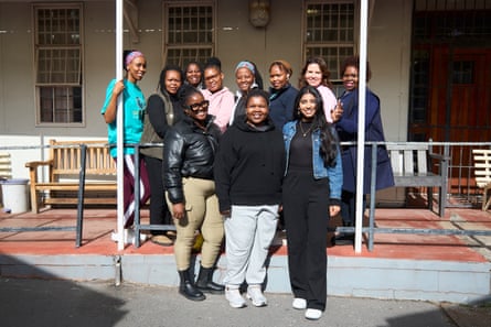 A group of smiling women stand on the veranda of a building in the sun