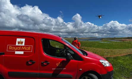 A postal worker standing beside a red Royal Mail van on a coast watches as a drone carrying a parcel takes off