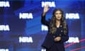 Woman with long brown hair waves on stage in front of NRA logo