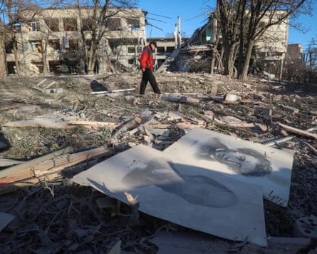 Man in red jacket walking through rubble with destroyed buildings in background and portraits on ground in foreground.