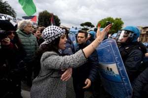 A woman in a headscarf holding up an arm and shouting at police, who are in riot gear with shields