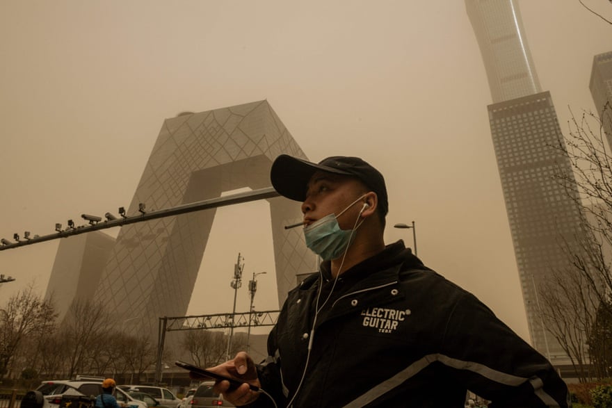 A man with a face mask around his chin smokes a cigarette with a dusty sky in the background