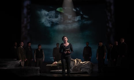 a woman (Lee Bisset playing Brunnhilde) stands singing in front of a group of people standing and one lying apparently dead on a hospital trolley in a dark, foreboding scene from Götterdämmerung at Longborough Festival Opera.