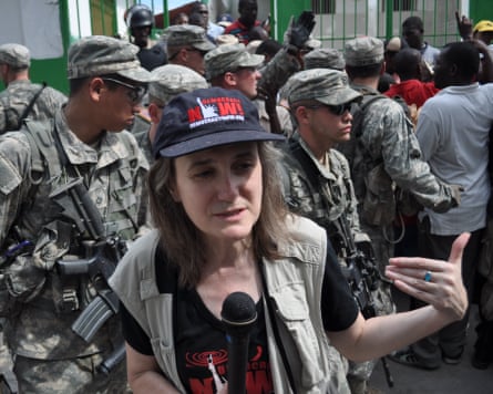 A woman with a baseball cap, khaki jacket and microphone in front of armed soldiers