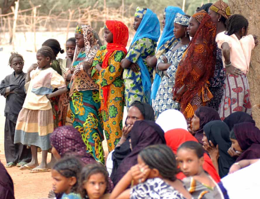 Black African girls, ranging in age from young children to teenagers, stand with grim expressions while paler African girls and women it in the foreground