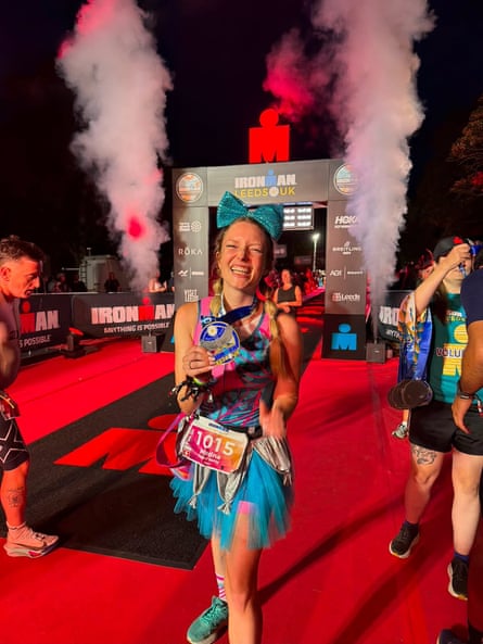 A smiling triathlete wearing a colourful costume and large blue bow holds a medal on the red finish carpet at an Ironman event in Leeds, with smoke rising behind her.