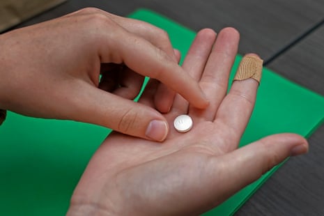 A young white right hand, palm up and a Band-Aid on the index finger, holds a white pill, while the left hand pinches as if to pick it up. Below is a table with a kelly green file folder.