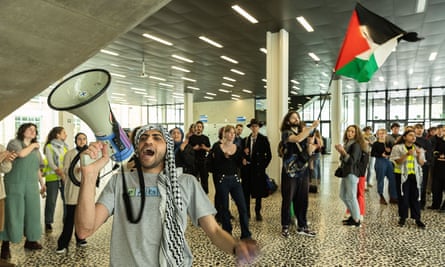 students gathered in foyer of university building; one is waving a Palestinian flag and another is shouting into a megaphone; some are wearing keffiyeh scarves 