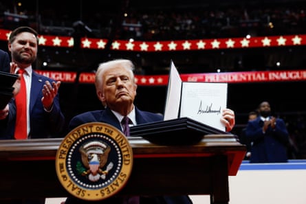 Donald Trump holds up a signed document in an arena setting with JD Vance clapping behind him