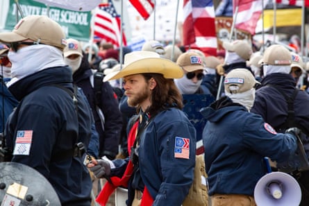 A man with a beard and long brown hair wearing a cowboy hat surrounded by members of the Patriot Front