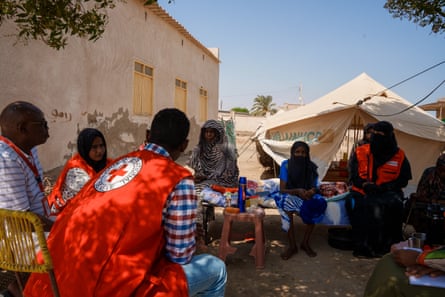 People, some wearing International Committee of the Red Cross red bibs, sit outside a relief tent.