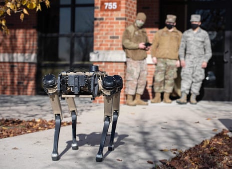 Three men in military fatigues watch in the background as a robot dog, a headless piece of technology that walks on four legs, stands on a leaf-strewn path.