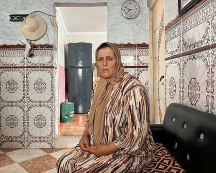An older Arab woman wearing a headscarf and djellaba robe sits in a tiled room