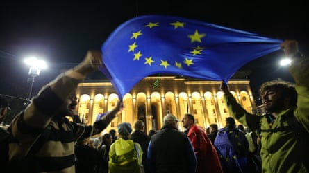 Demonstrators wave an EU flag in front of the Georgian parliament building in Tbilisi: the building is seen illuminated under the flag, which is held by two young men either side; it is dark, with streetlights glowing, and a crowd is seen gathered in front of the two men; people are wearing coats and waterproof capes, and one person is seen with another EU flag draped over their shoulders.
