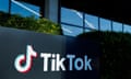 Black sign with TikTok logo in front of bushes and building windows