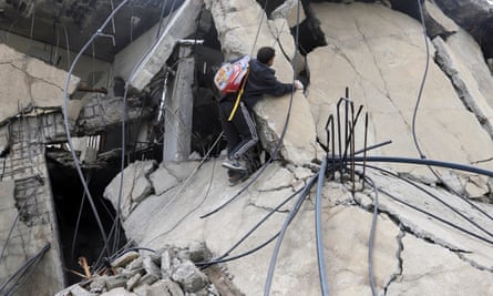 A Palestinian man wearing a child’s backpack climbs the damaged structure of a building hit by the Israeli airstrike; there are wires, pipes and chunks of concrete around him.
