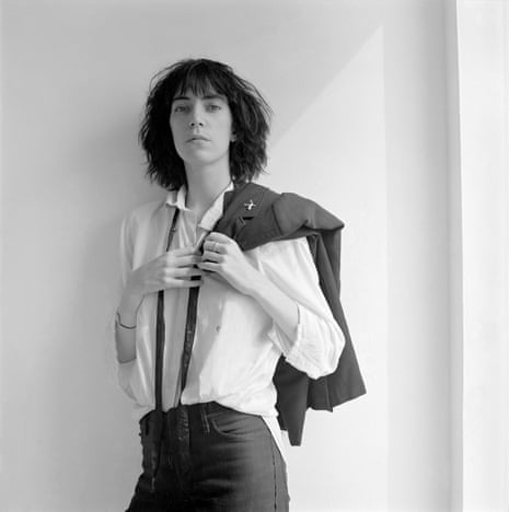 Musician Patti Smith, in white shirt and black suit, leaning against a white wall