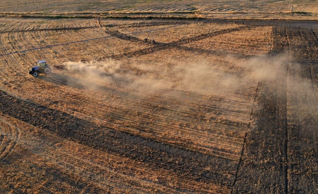 An aerial view of a dry field.  A tractor plowing the field produces a long cloud of dust that carries the wind.